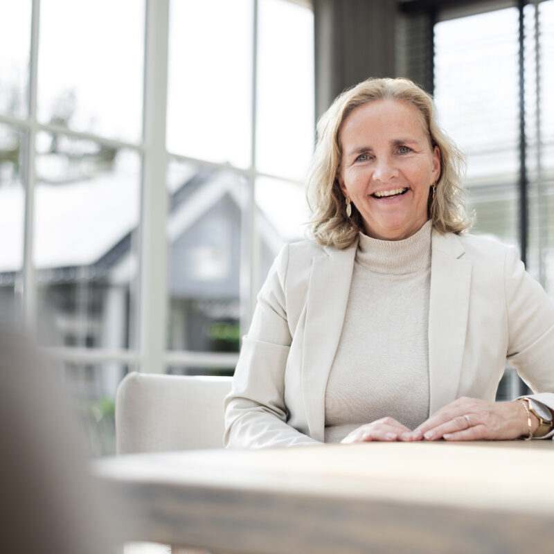 Annelies in een lichte ruimte, ontspannen en vriendelijk aan tafel.