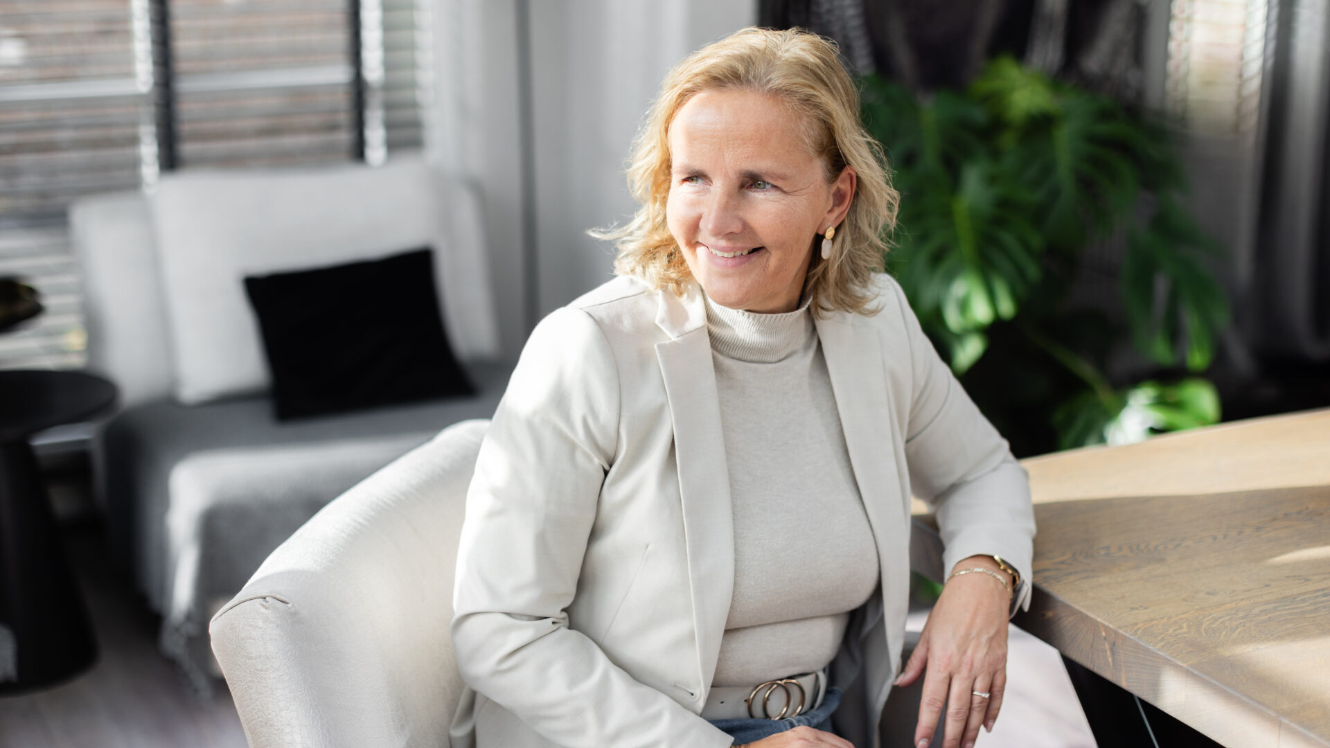 Coach in lichte werkruimte, glimlachend aan tafel tijdens een coachingsgesprek of reflectiemoment.