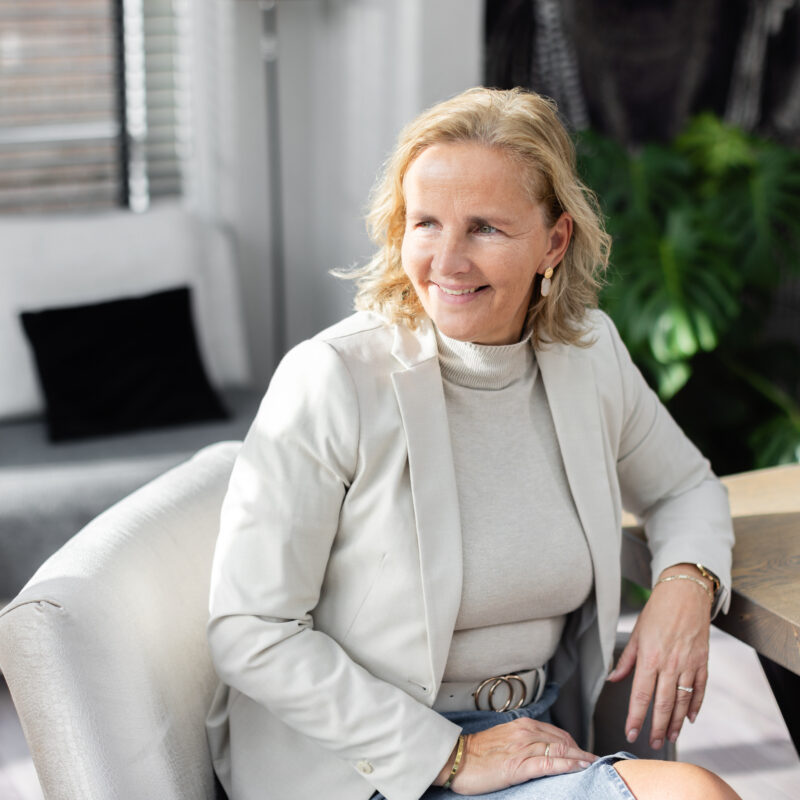 Coach in lichte werkruimte, glimlachend aan tafel tijdens een coachingsgesprek of reflectiemoment.