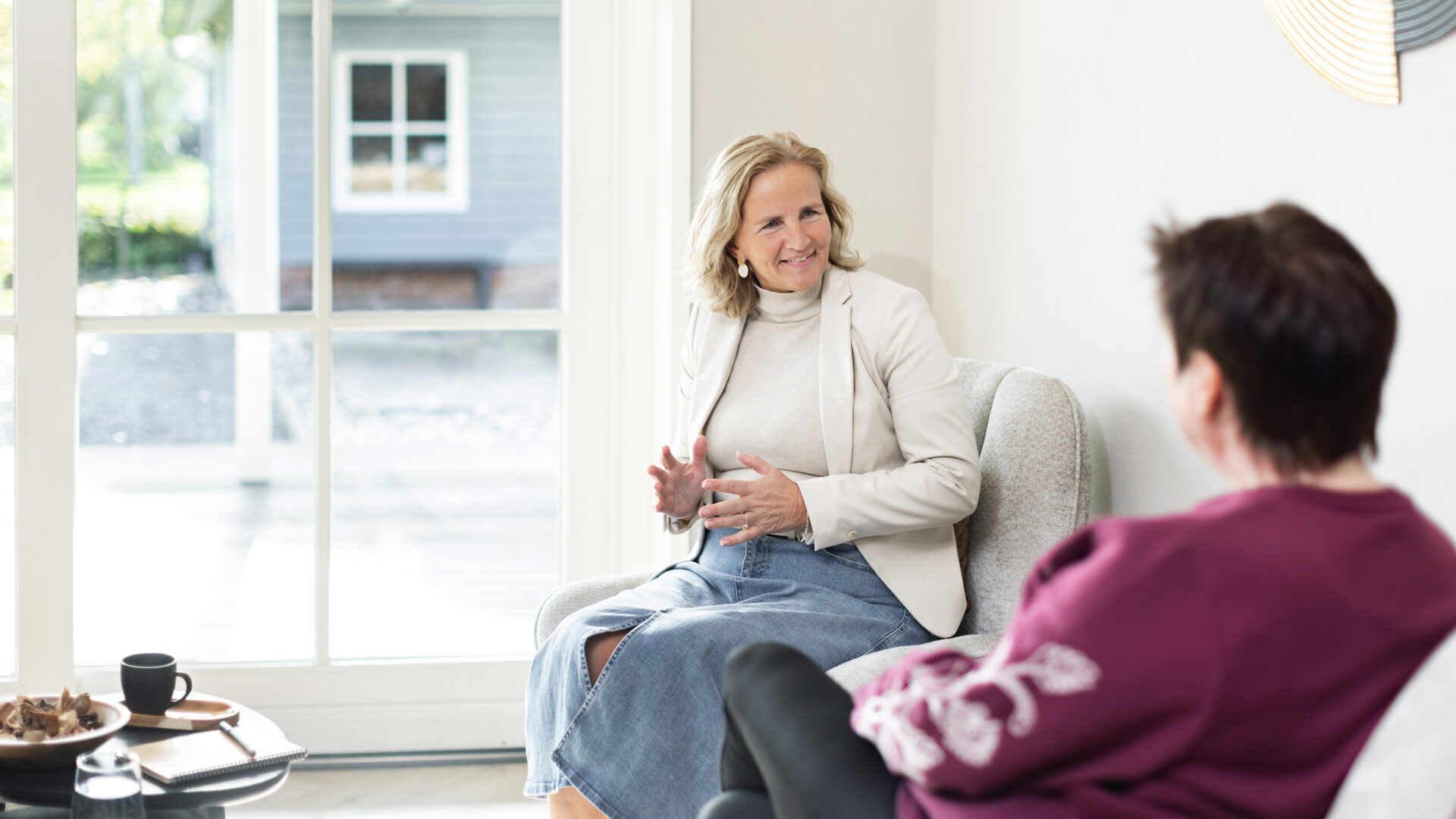 Annelies in gesprek met een bezoeker in een lichte coachingruimte met comfortabele fauteuils.